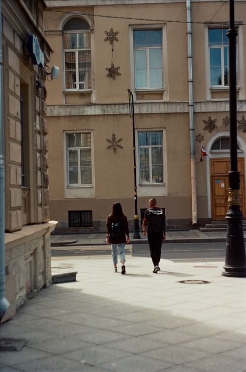 Free A couple walks down a sunlit urban street, showcasing city architecture and casual outdoor lifestyle. Stock Photo