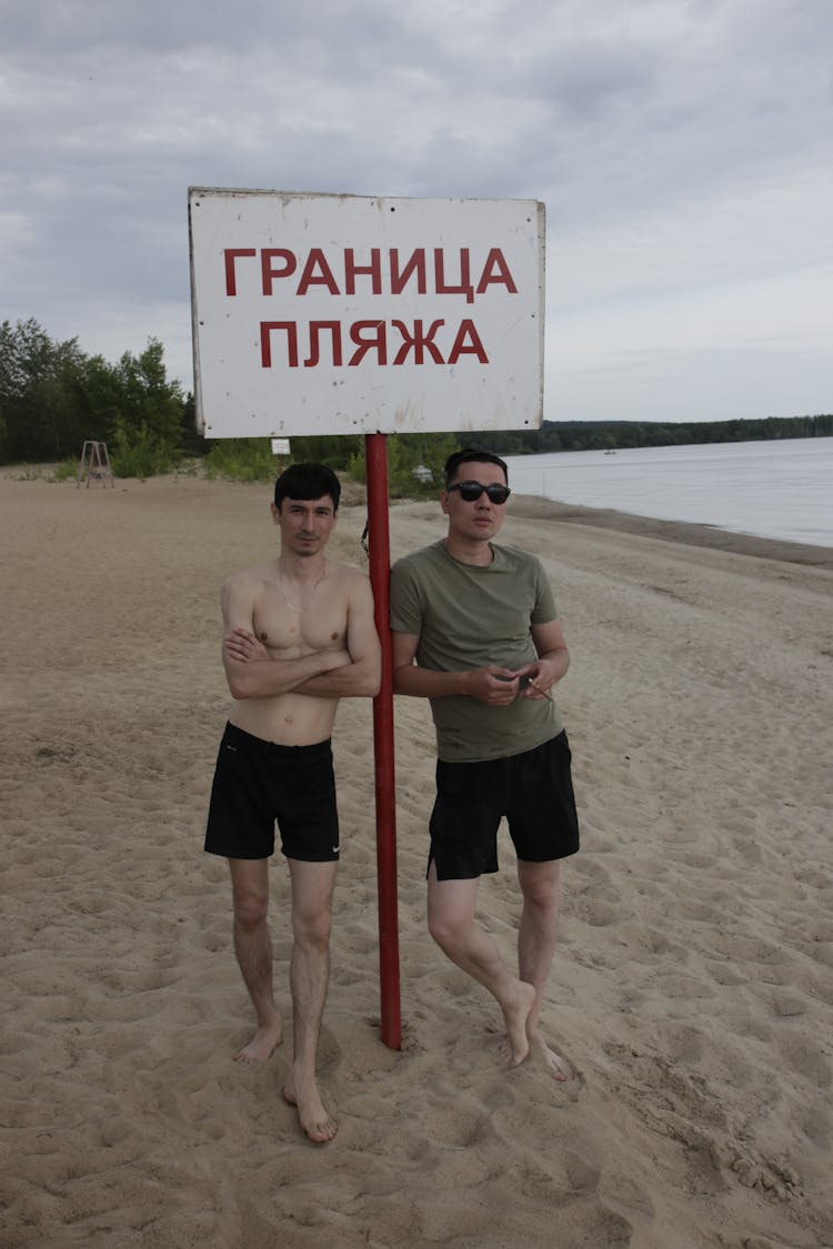 Two Men Standing At The Beach Beside The Signpost