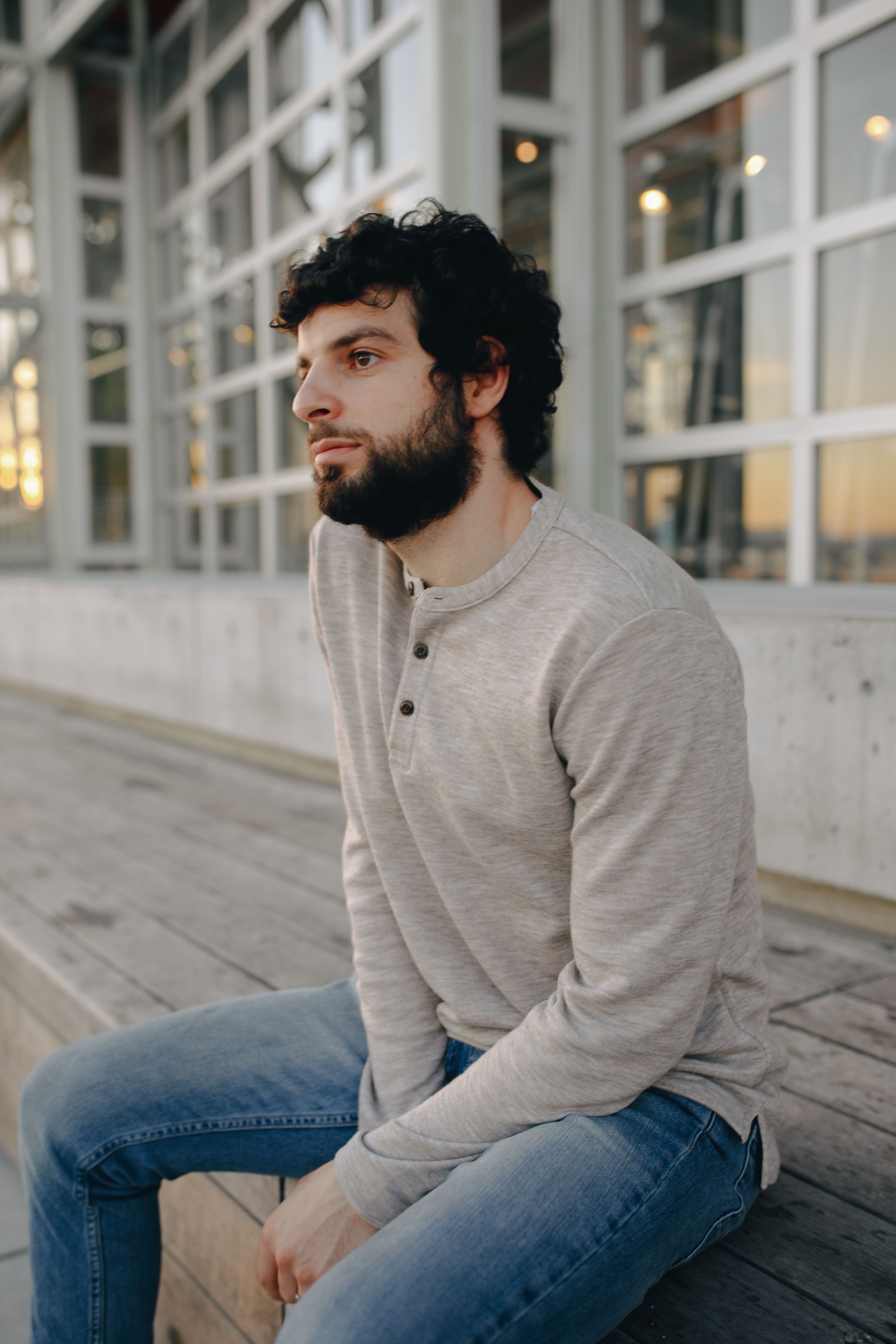 A man with curly hair and beard sitting on wooden steps wearing a gray sweater and jeans.
