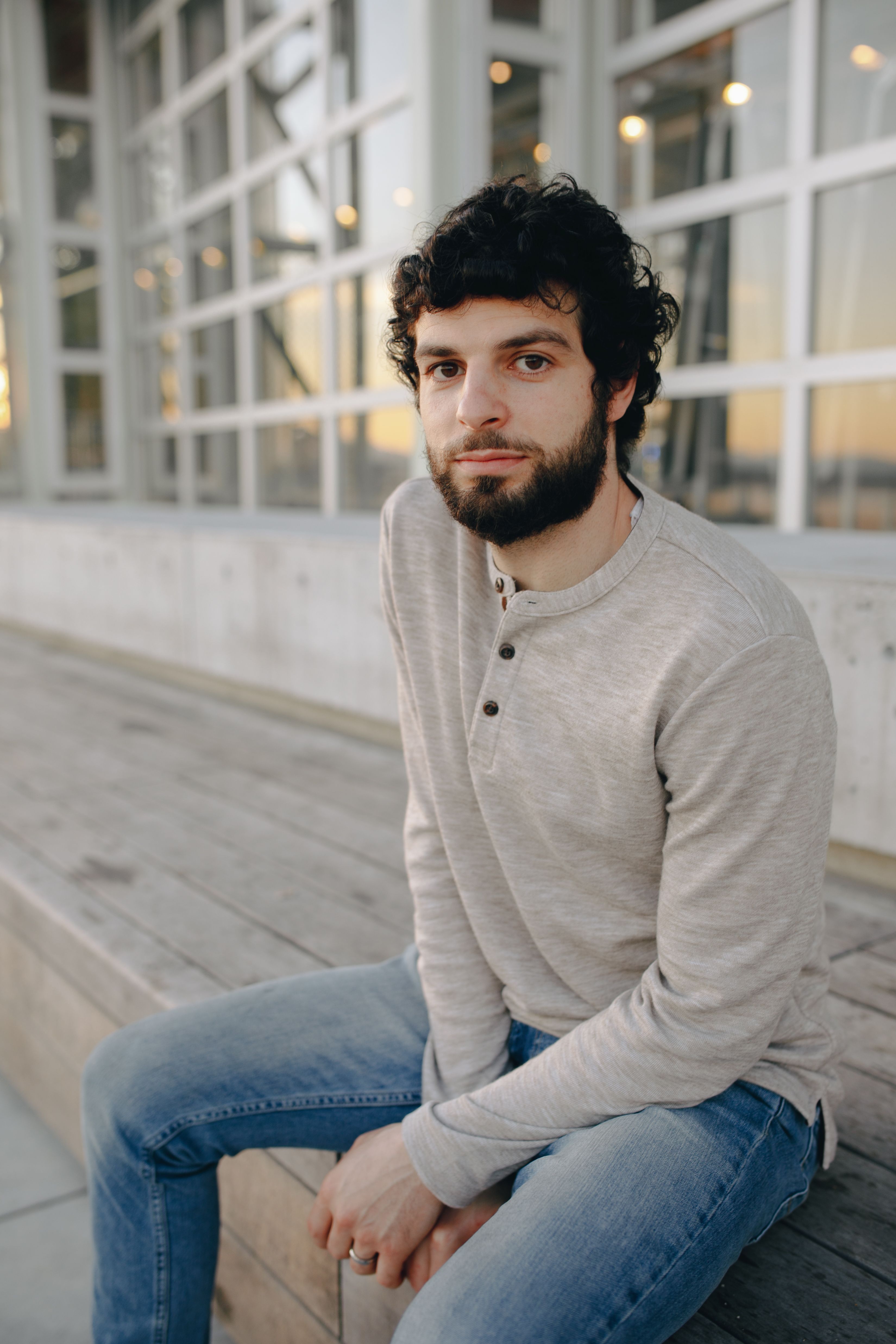 A bearded man in a gray sweater sitting on wooden steps outdoor setting.