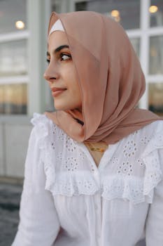 Portrait of a woman in a peach hijab and white lace blouse, elegantly styled, gazing thoughtfully.