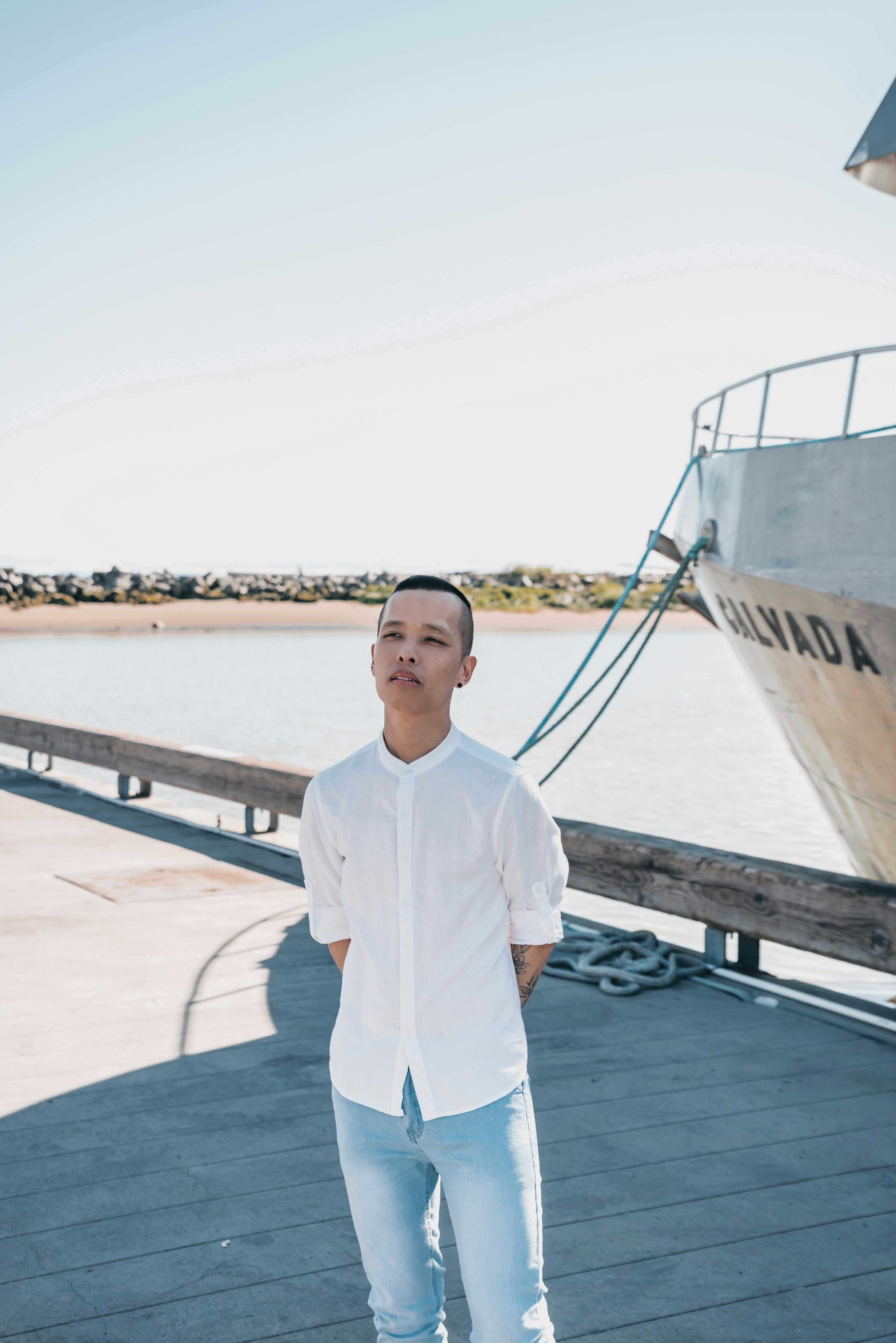 Young man standing on a dock in sunlight, with a boat and water in the background.