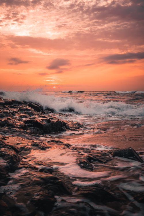 Free Waves crash against a rocky coast during a vivid sunset, creating a dramatic seascape. Stock Photo