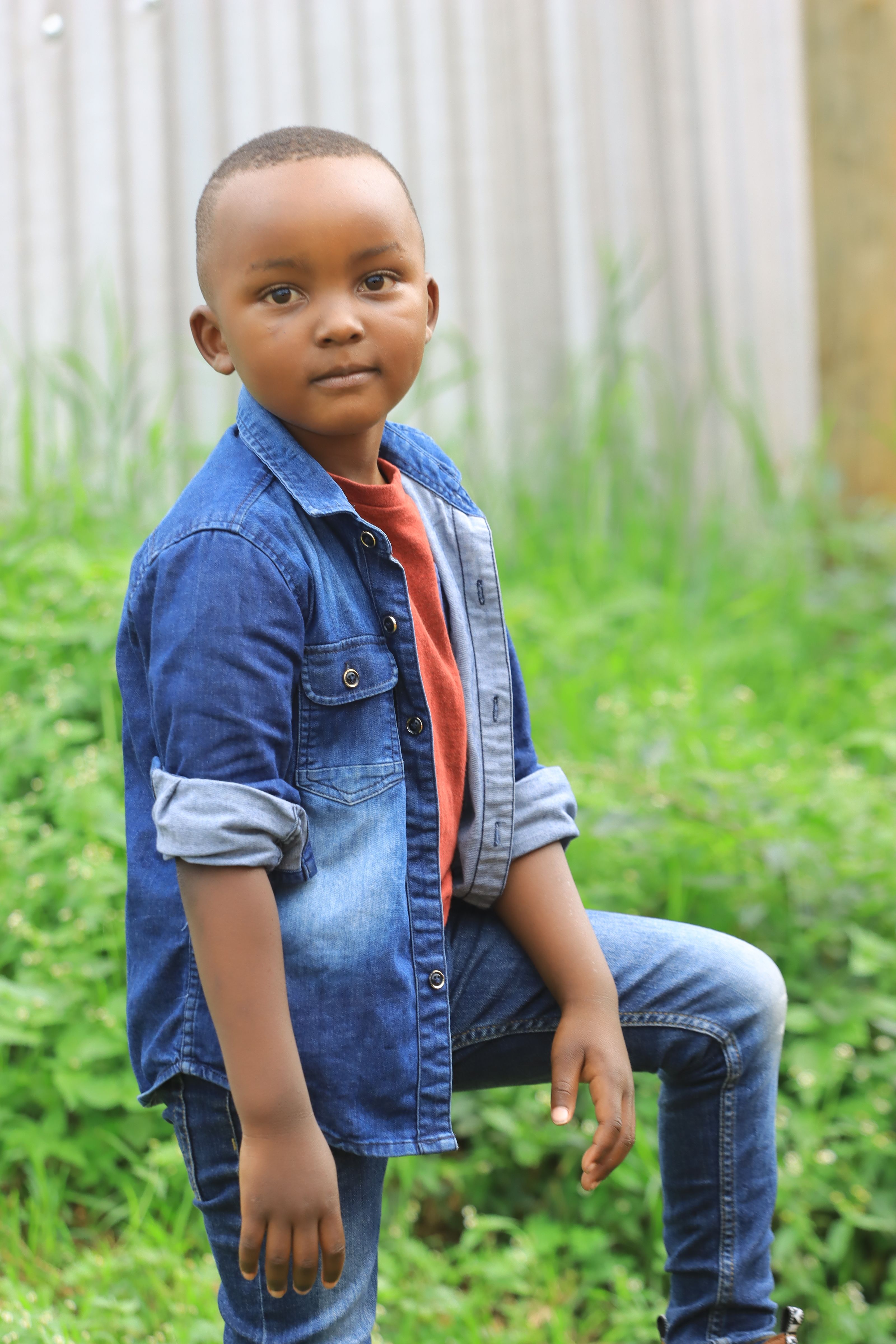 Young boy posing confidently outdoors in casual denim outfit, against a backdrop of lush green grass.