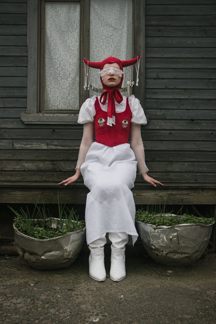 Woman In Eccentric Costume Sitting On Bench Near Wooden House