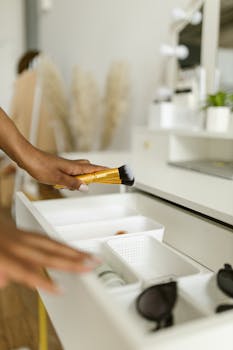 A person holding a makeup brush organizing cosmetics in a white drawer.