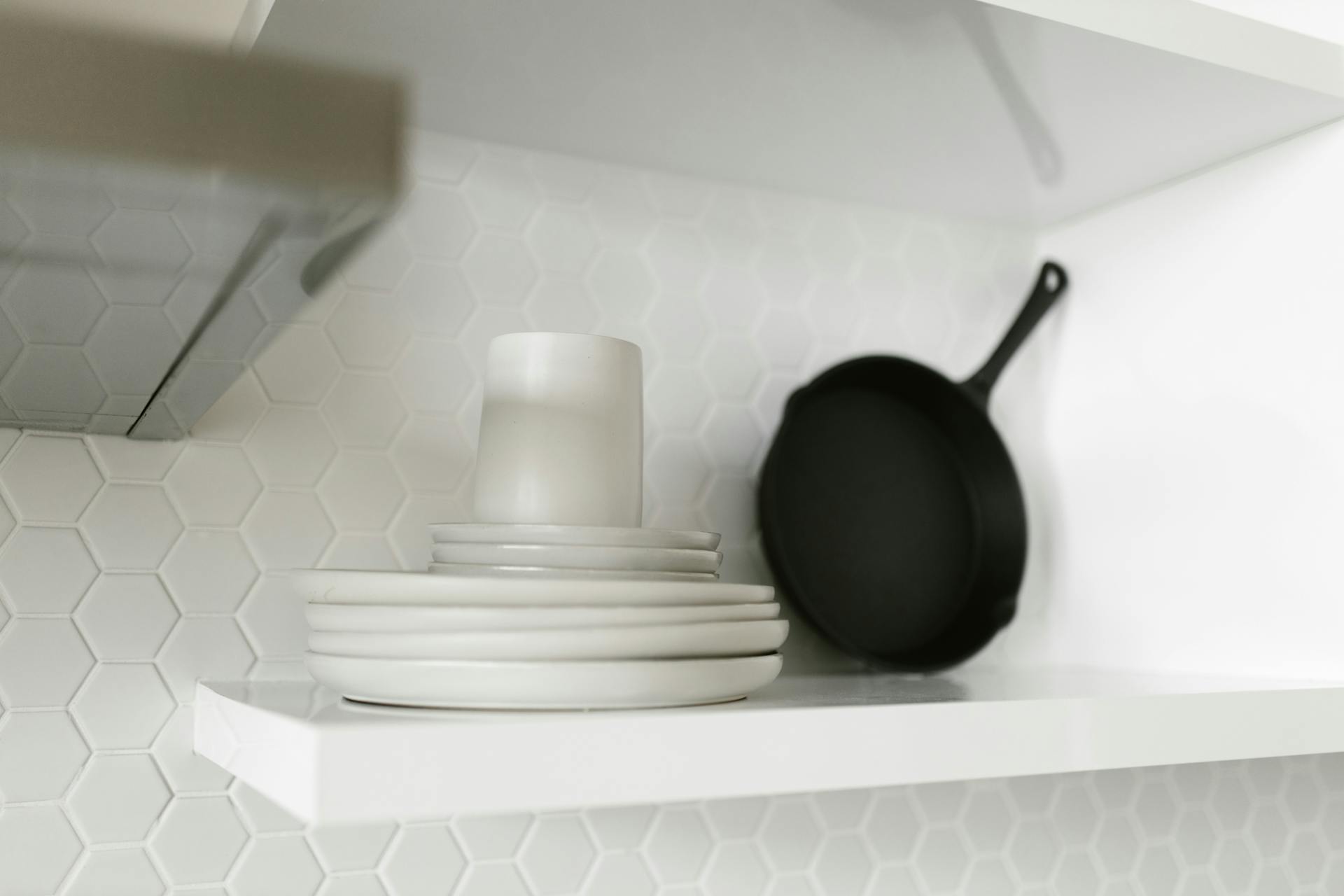 A minimalist white shelf displaying stacked plates and a black pan against a hexagonal tiled wall.