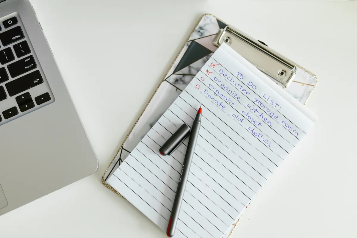 A clipboard holding a handwritten to-do list with two items checked off, a red-tipped pen and a laptop at the edge of a white desk.