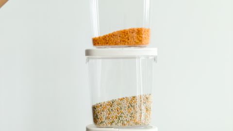 Hands holding stacked plastic containers with various dry foods against a neutral background.