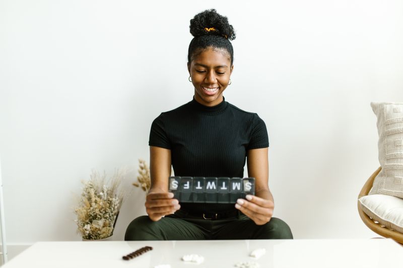 A smiling young woman holds a labeled weekly pill organizer at her table, a few pills laid out in front of her.