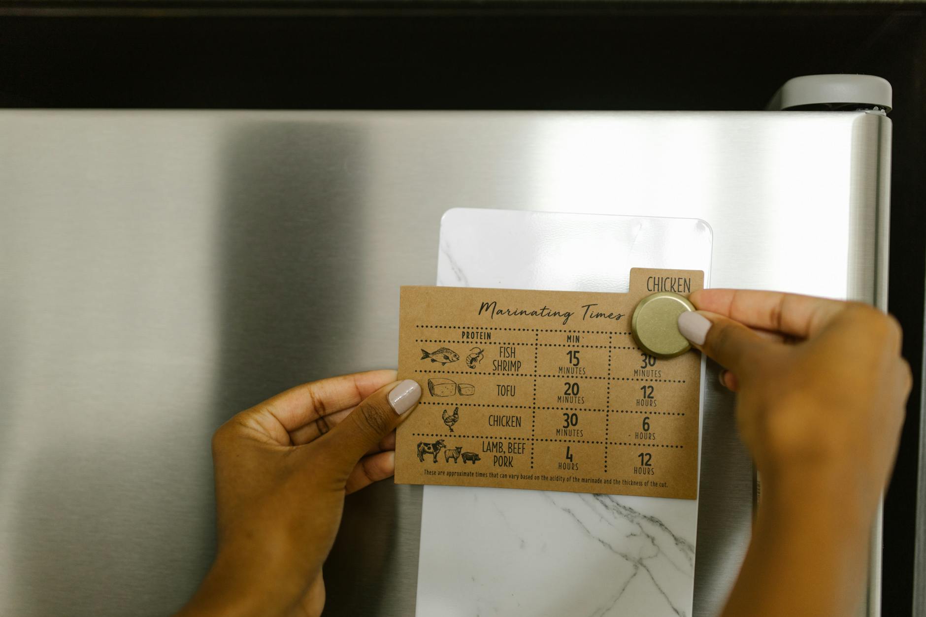 Hands placing a marinating timer card on a fridge with a magnet, featuring cooking times for various proteins.