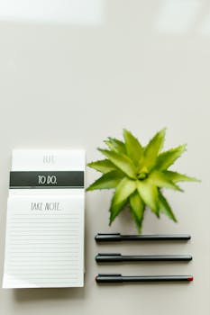 Elegant flat lay of a notepad and pen beside a green plant on a clean white surface.
