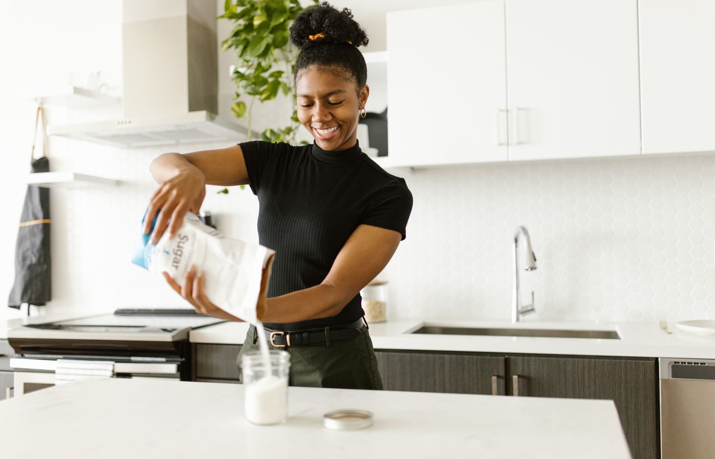 Smiling woman pouring sugar into a glass container in a bright, modern kitchen setting.