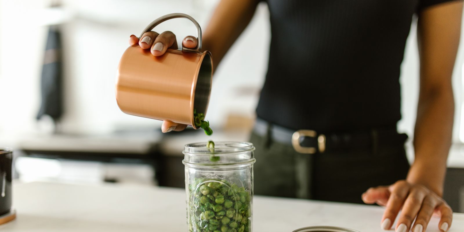 A woman pours green peas into a glass jar on a kitchen counter, emphasizing organization.