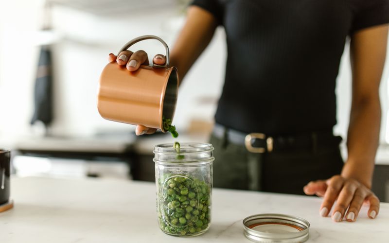 A woman pours green peas into a glass jar on a kitchen counter, emphasizing organization.