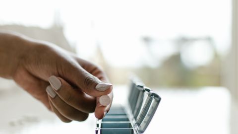 A close-up of a hand organizing pills in a weekly pill container on a bright surface.