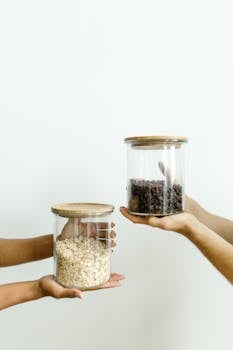 Two hands hold glass jars filled with grains and beans against a white wall.
