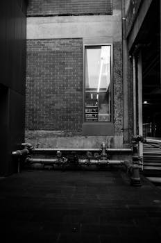 Black and white image of an urban industrial scene featuring a brick wall and metal pipes.