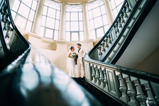 A bride and groom elegantly pose on a grand staircase in a classic indoor setting.
