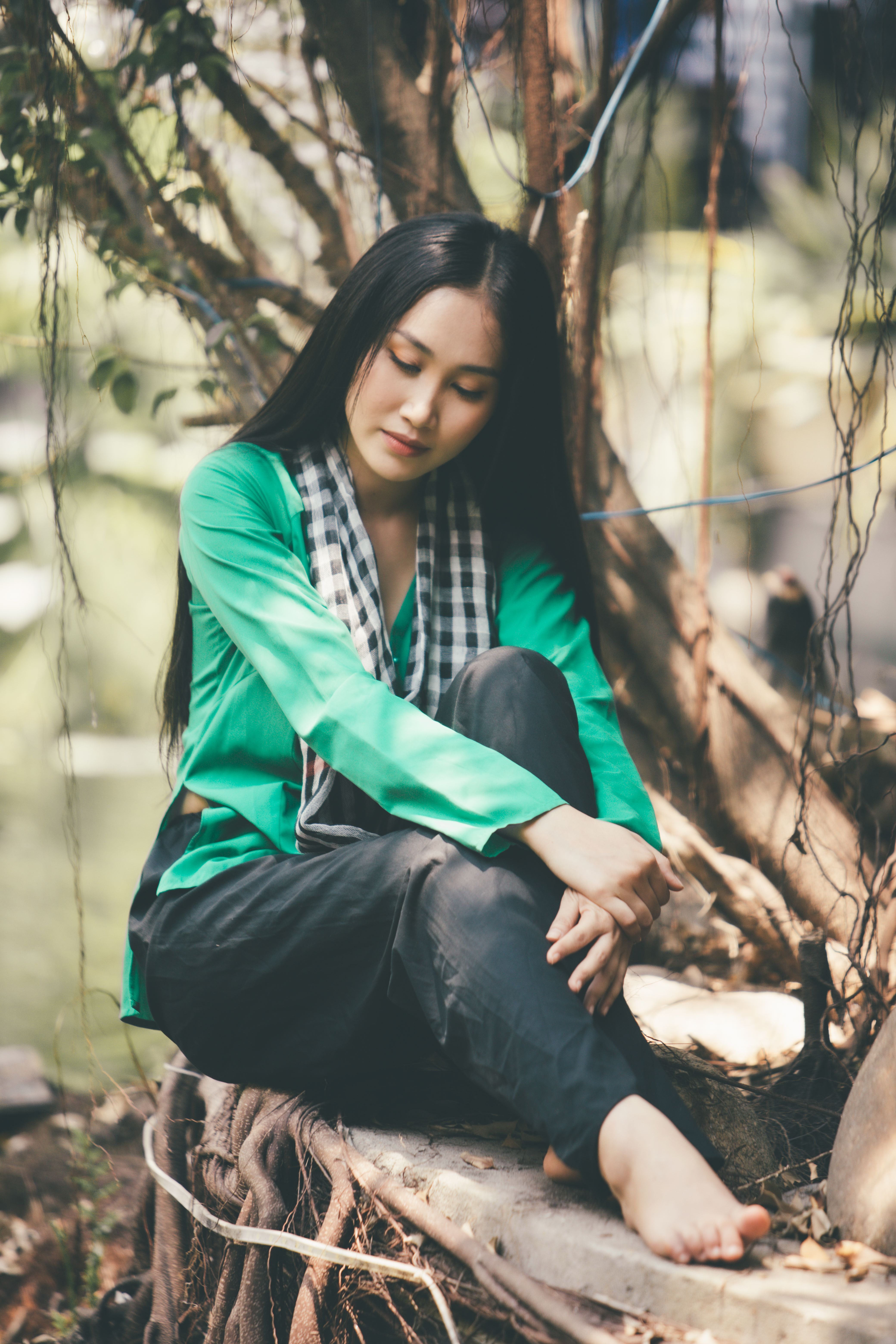 Asian woman in green shirt and scarf sitting outdoors, barefoot on a tree, serene and peaceful.