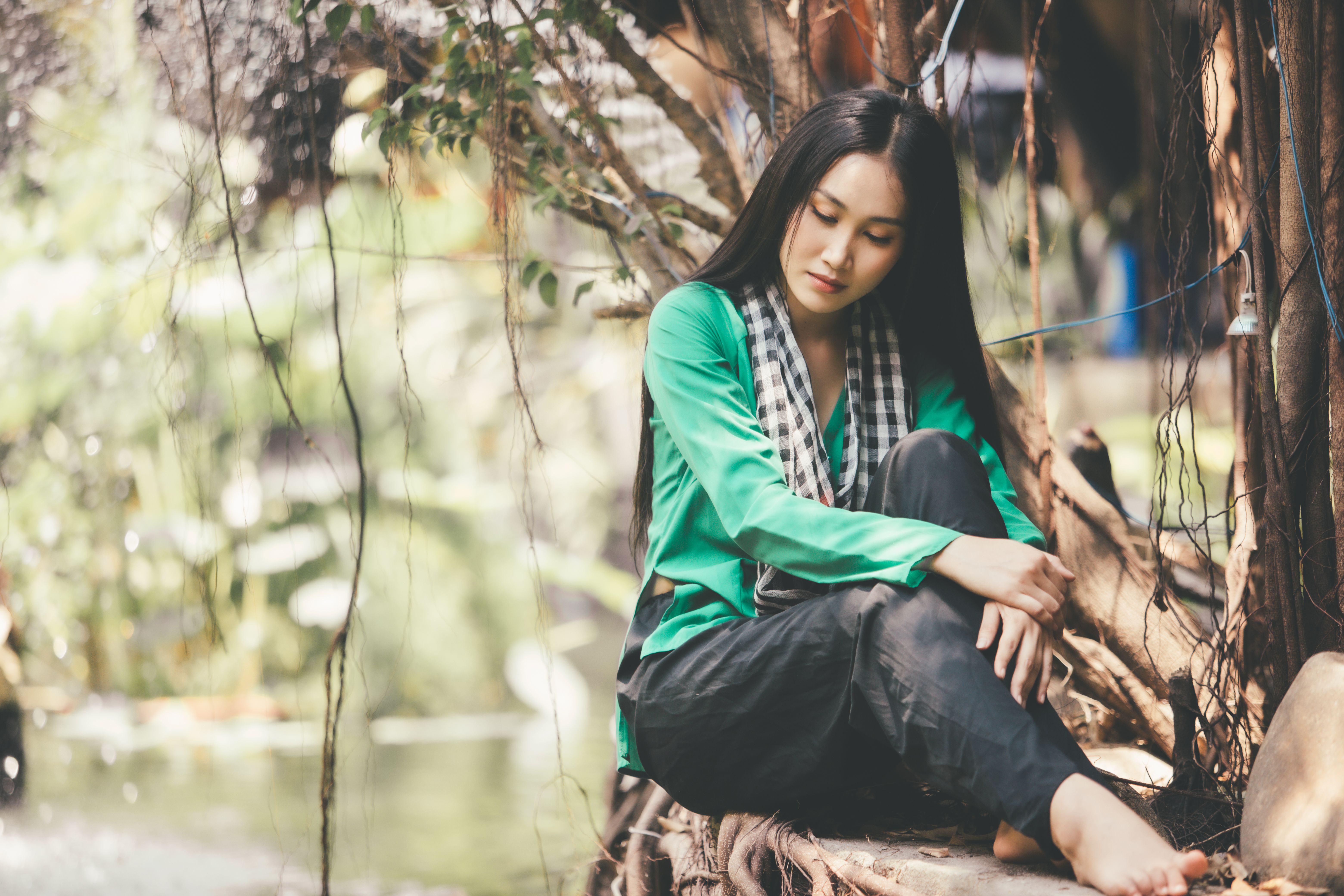 Asian woman in green shirt sitting barefoot by a tree, enjoying a peaceful outdoor setting.