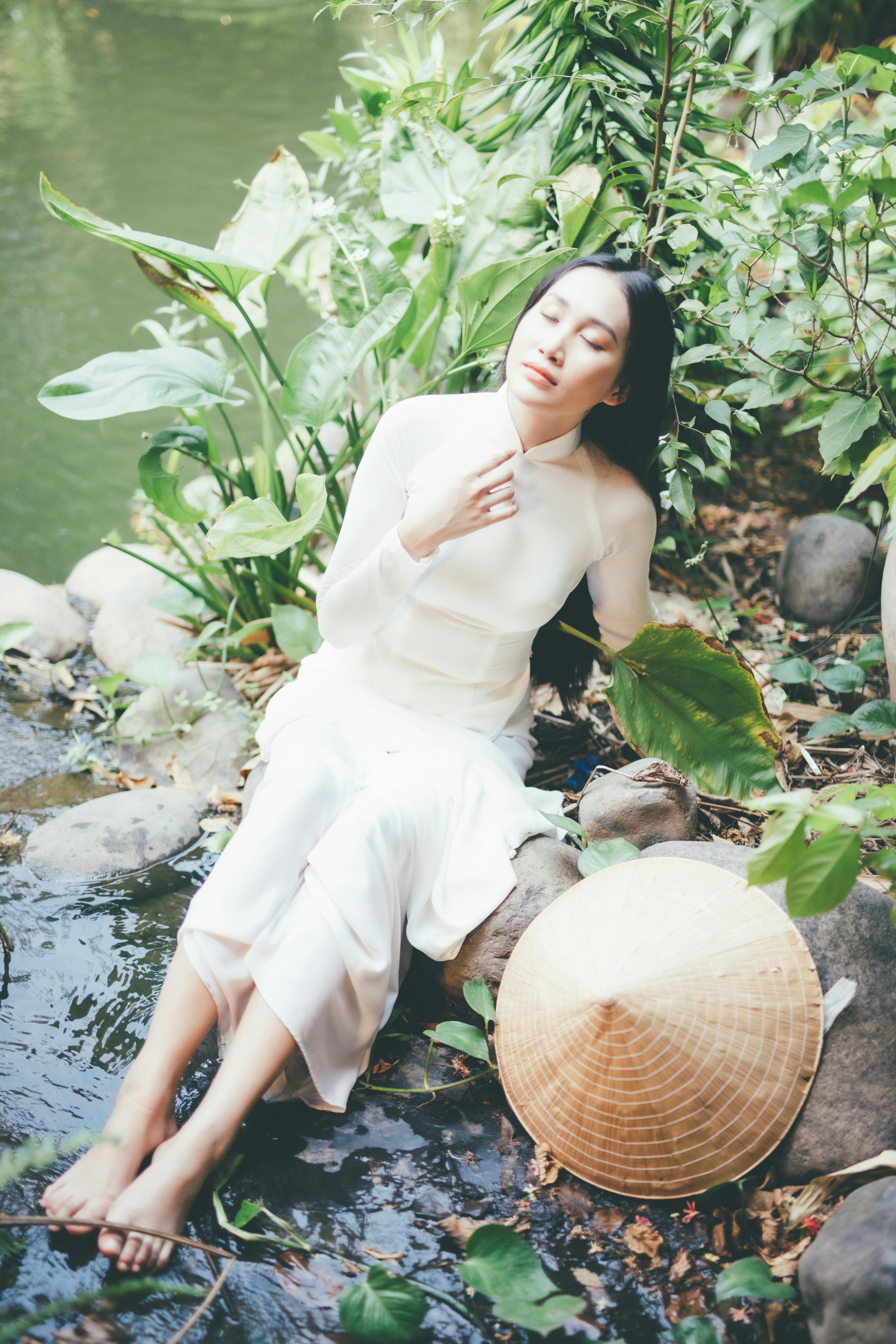 An elegant woman in a white dress sits peacefully by a tranquil river surrounded by lush greenery.