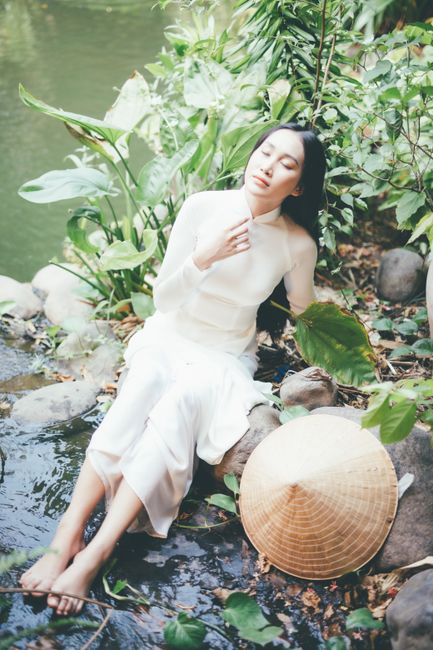 An elegant woman in a white dress sits peacefully by a tranquil river surrounded by lush greenery.
