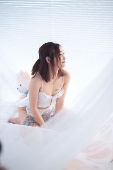 An Asian woman wearing lingerie while kneeling on a bed, illuminated by soft daylight.