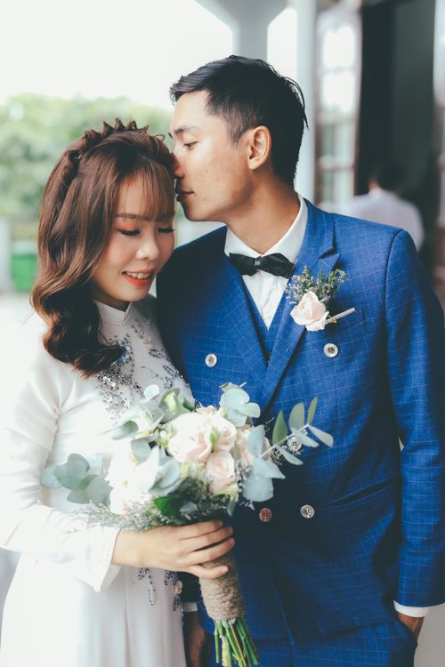 Free Asian couple celebrating their wedding day outdoors, sharing a tender moment with a bouquet. Stock Photo