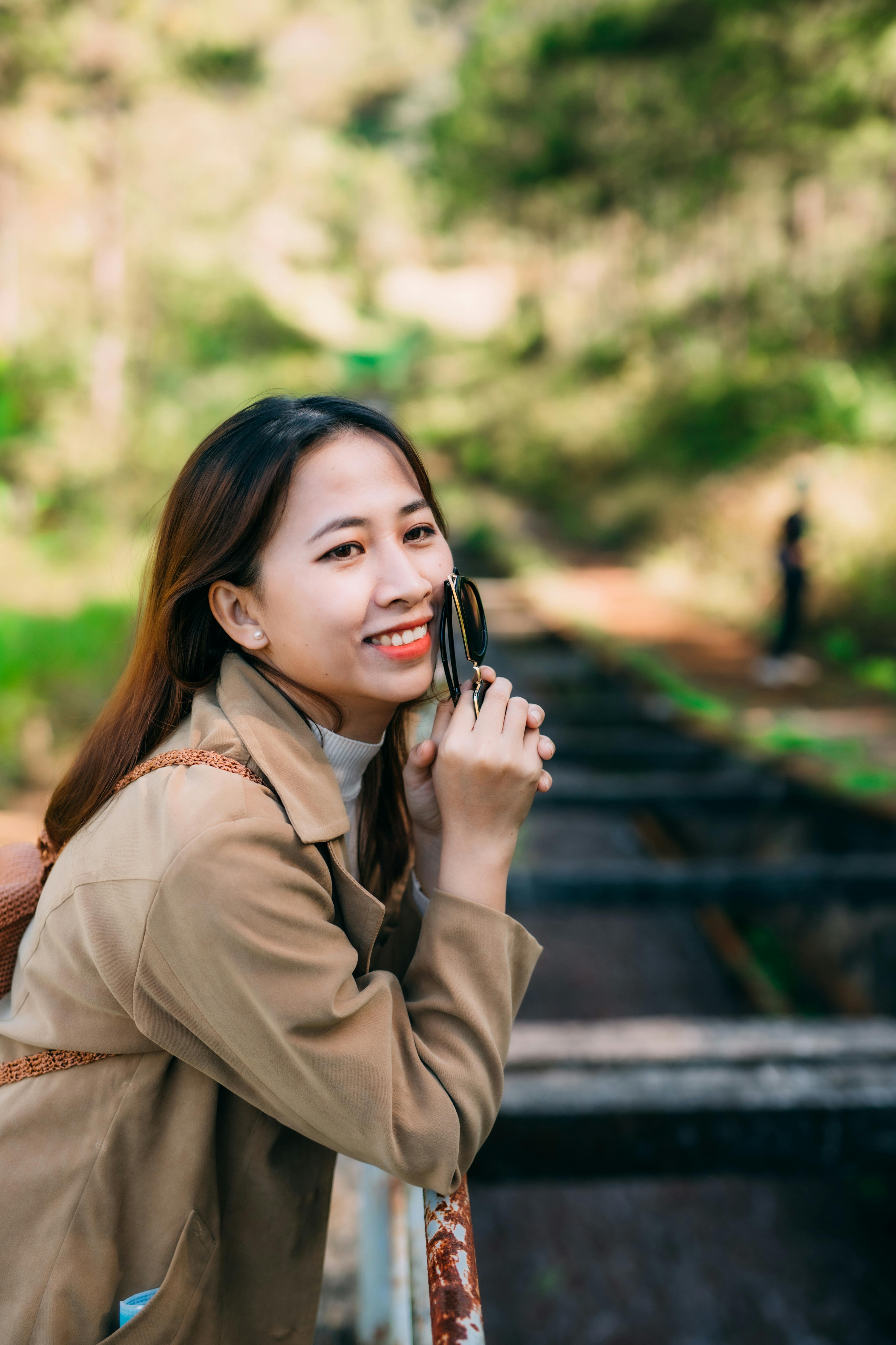 A joyful woman in a brown jacket smiling while enjoying the natural surroundings during a sunny day.