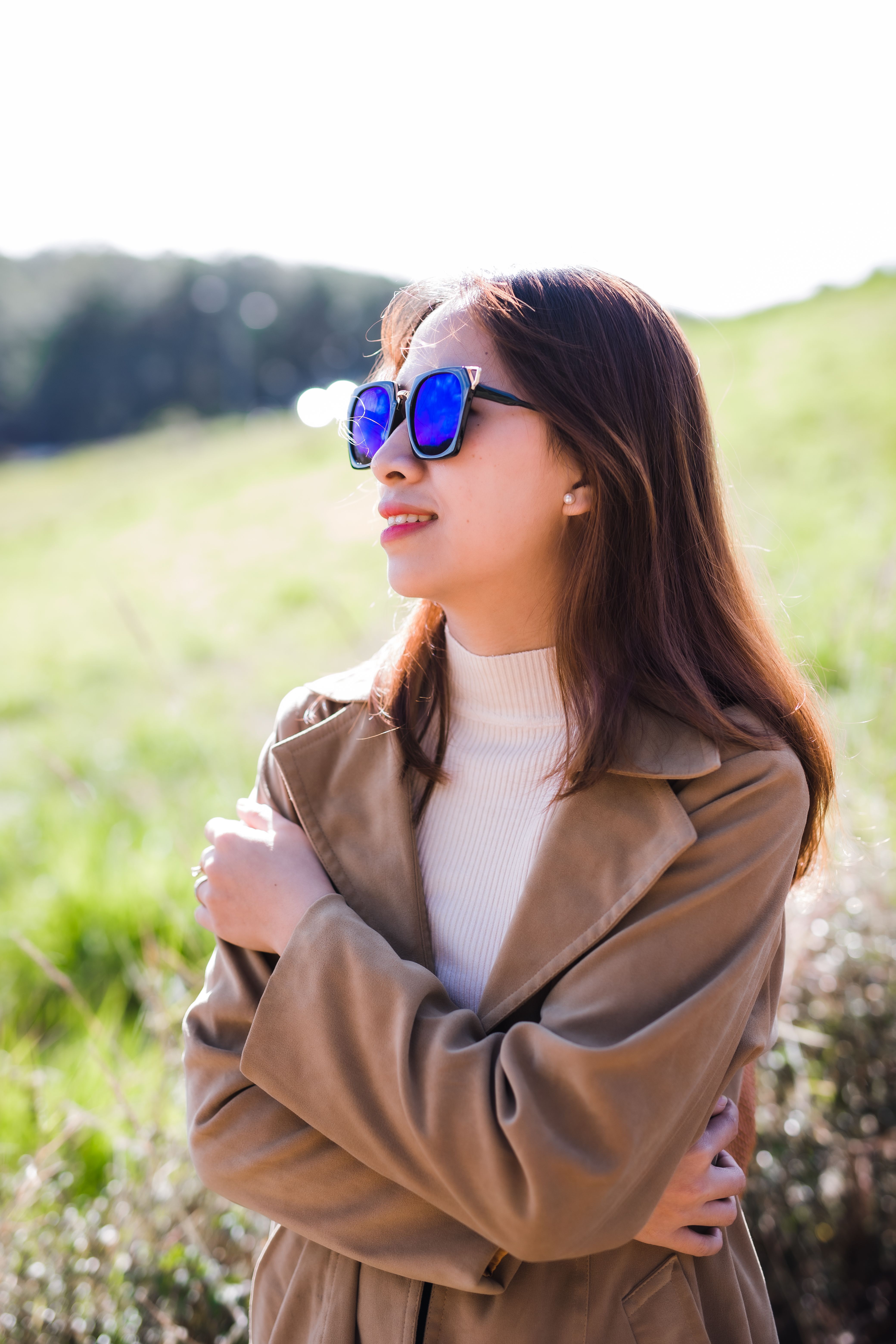 A fashionable young woman in sunglasses enjoying a sunny day outdoors, wearing a brown jacket.