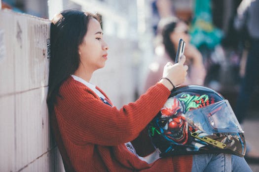 Asian woman sitting outdoors, wearing knitted sweater, checking phone with vibrant helmet beside her.