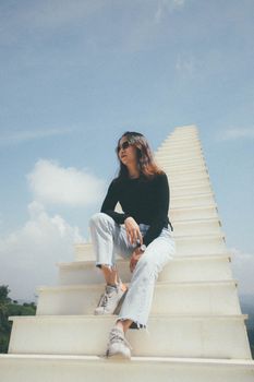 A woman in black long sleeves sits stylishly on a white staircase under a blue sky.