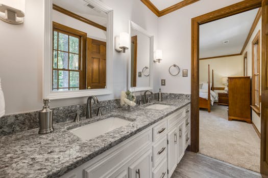 A stylish washroom with granite countertop, double sinks, and view into a bedroom.