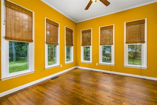 Spacious room with wooden floors, yellow walls, and large windows overlooking greenery.
