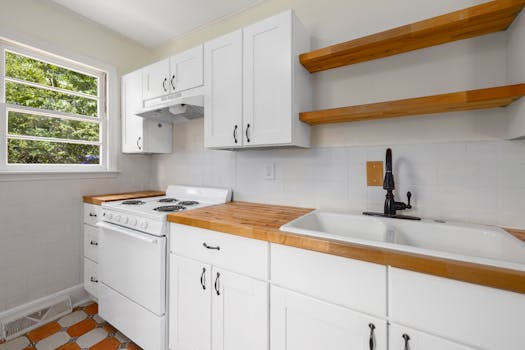 A bright kitchen with white cabinets, wooden shelves, and a large window.