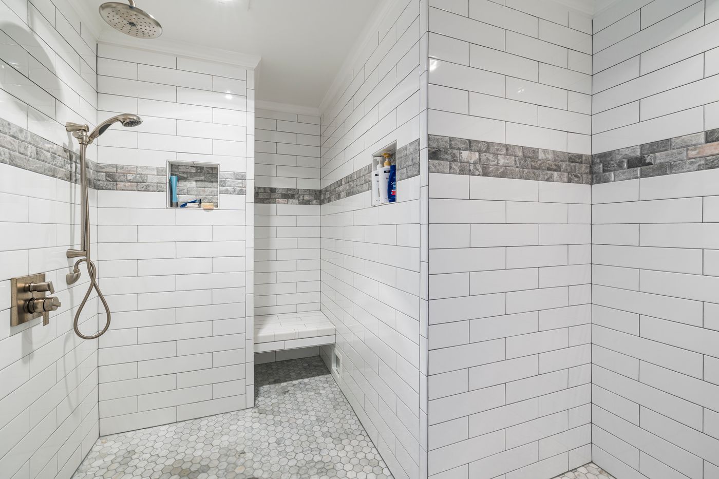 Large walk-in shower clad in white subway tiles, with a contrasting stone-look accent stripe, built-in niches, bench, handheld shower and rain shower head.