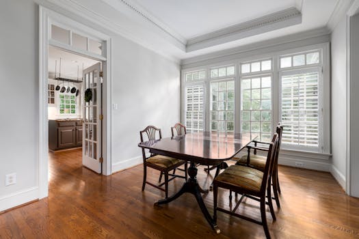 Bright dining room featuring a wooden table, classic chairs, and large windows overlooking greenery.