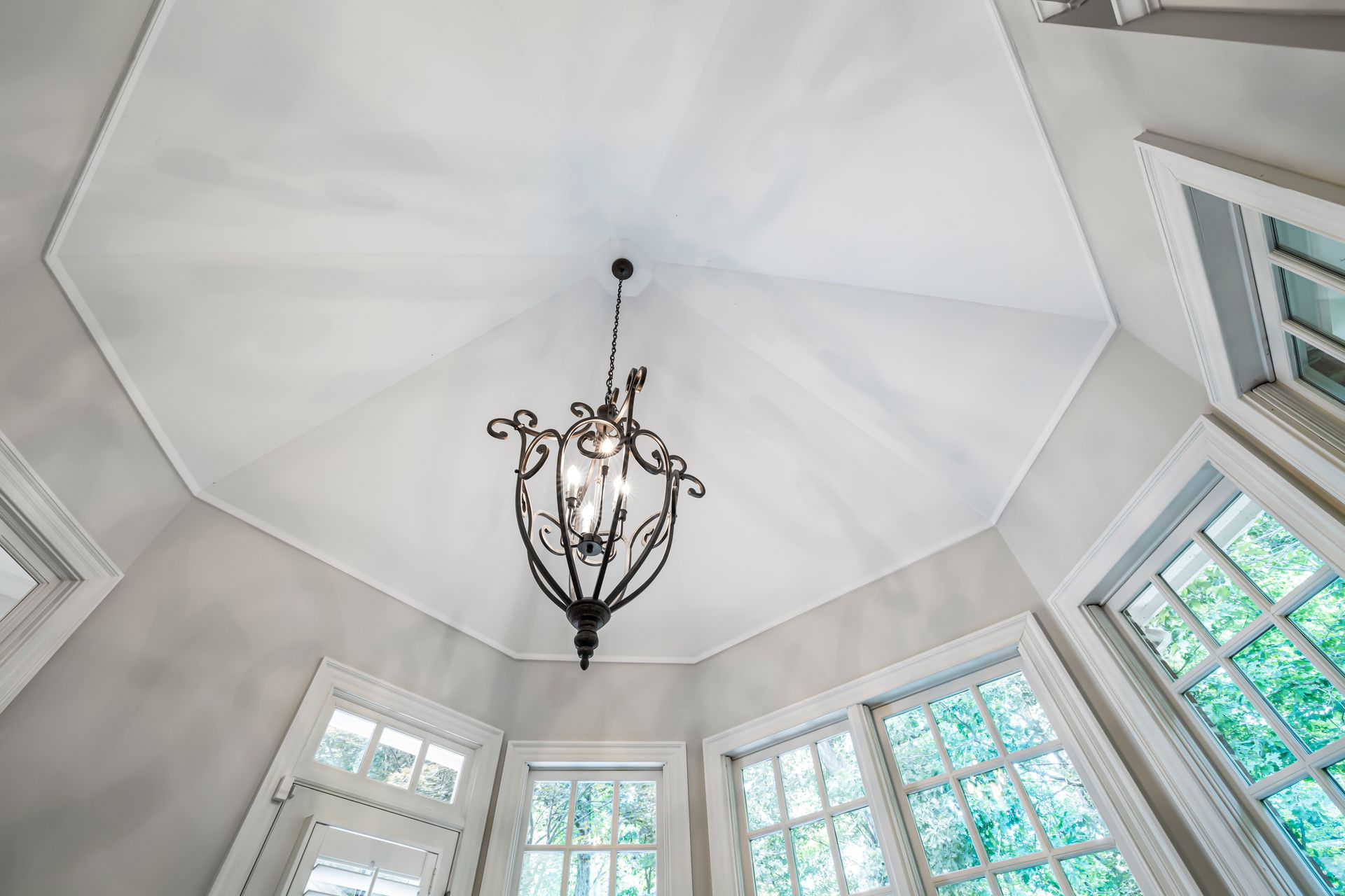 A wrought iron chandelier hanging in a sunlit room with large glass windows and white ceiling.
