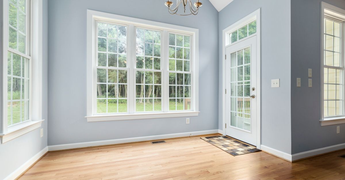 Sunlit empty room with wooden floor, chandelier, and French windows leading outside.
