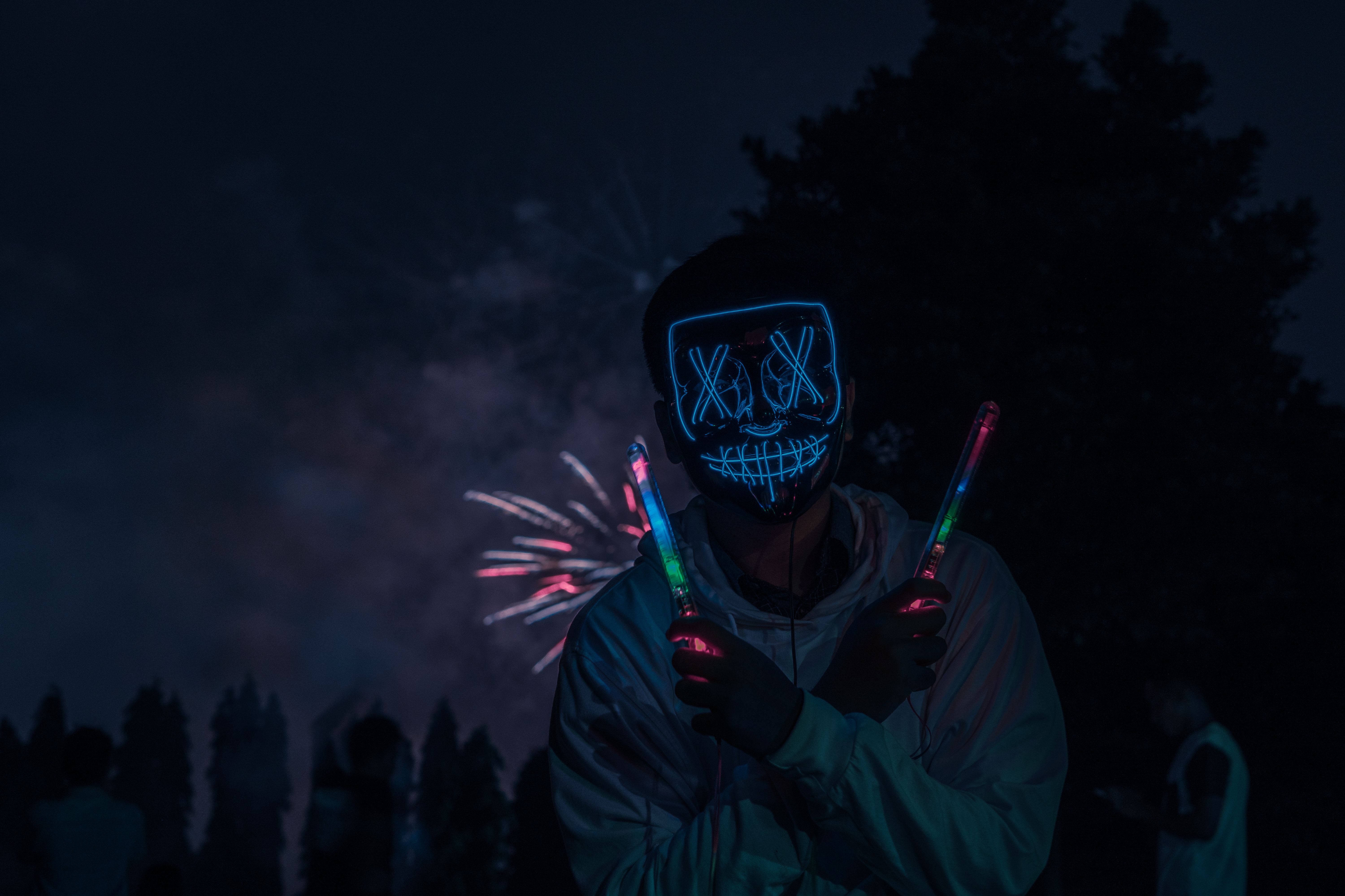 A person wearing a neon mask holding glow sticks with fireworks in the background, creating an illuminated night scene.