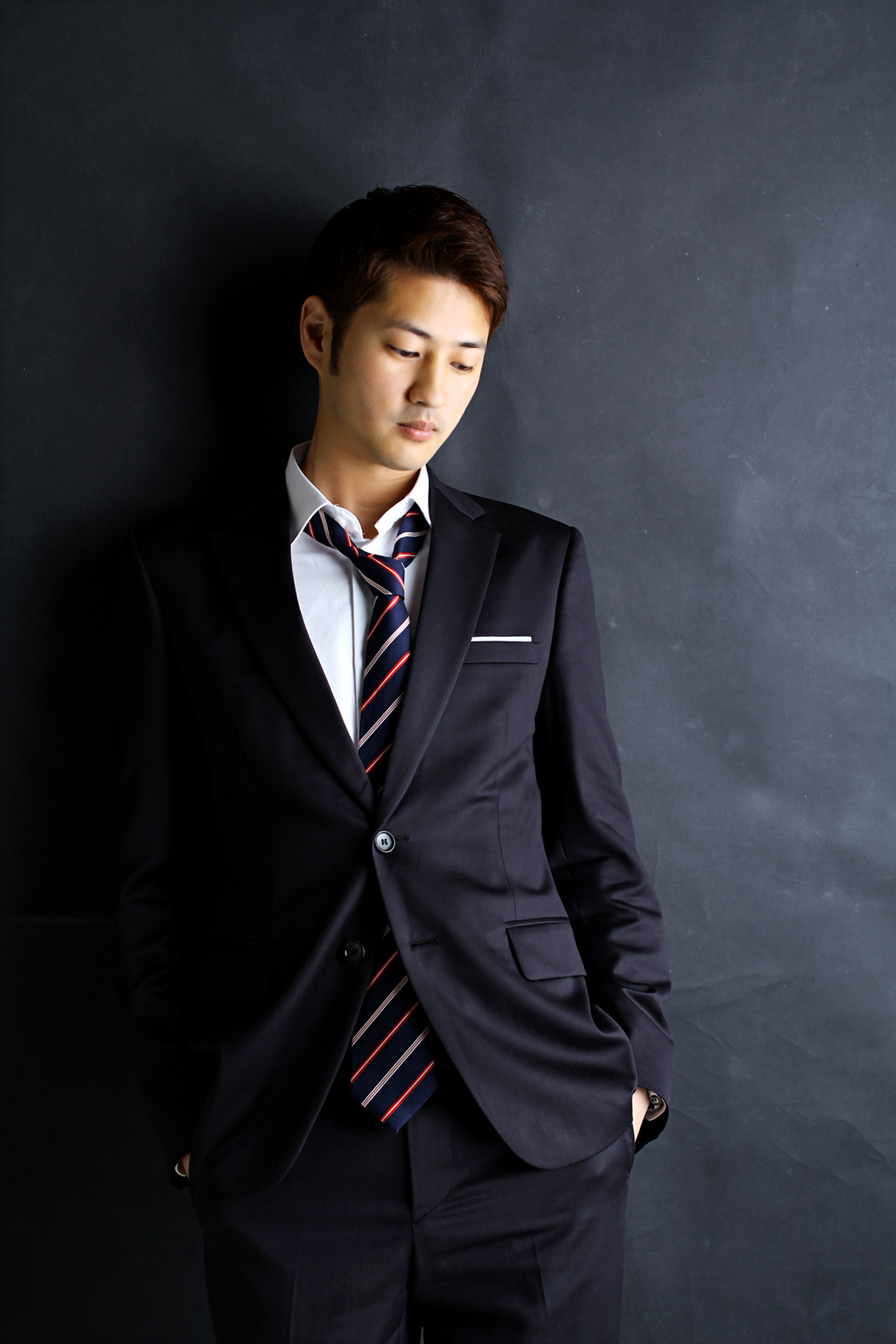 Stylish young man in a black suit poses confidently indoors against a dark background.