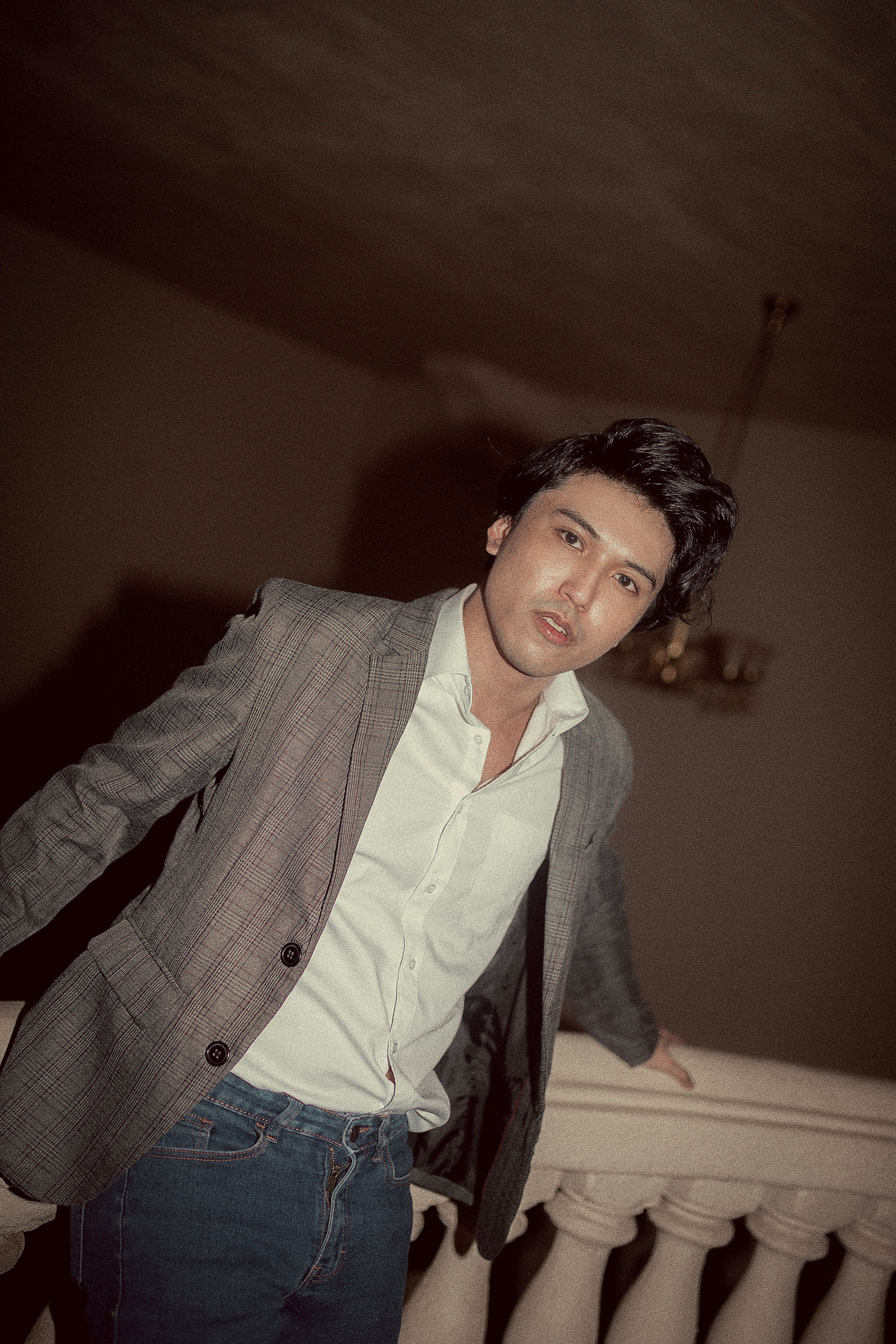 Young man in a gray blazer and jeans leaning on a railing indoors.