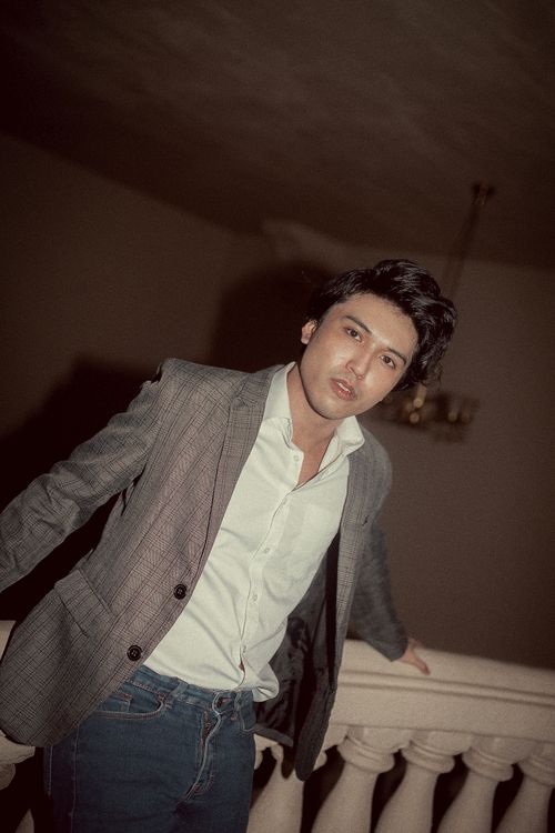 Free Young man in a gray blazer and jeans leaning on a railing indoors. Stock Photo