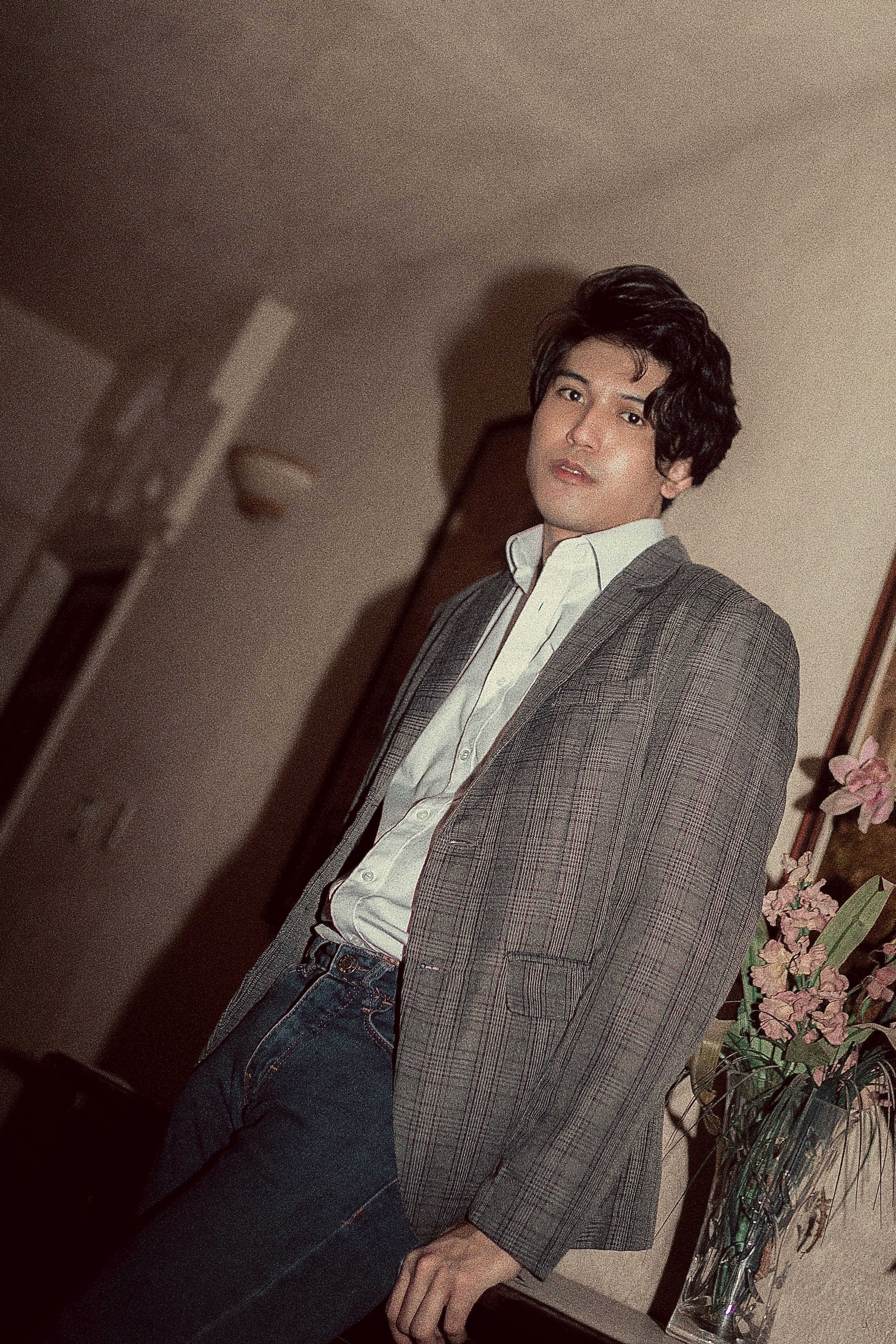 Man in a blazer and jeans leaning stylishly near flowers indoors.