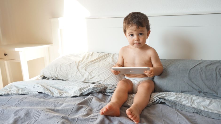 How to Choose Between Cloth and Disposable Diapers: Cost & Eco Impact A cute baby sitting on bed with tablet, enjoying a relaxed indoor moment.