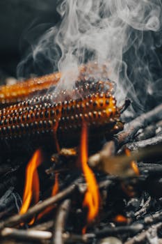 Close-up of corn grilled over open flames with rising smoke, capturing the rustic cooking ambiance.