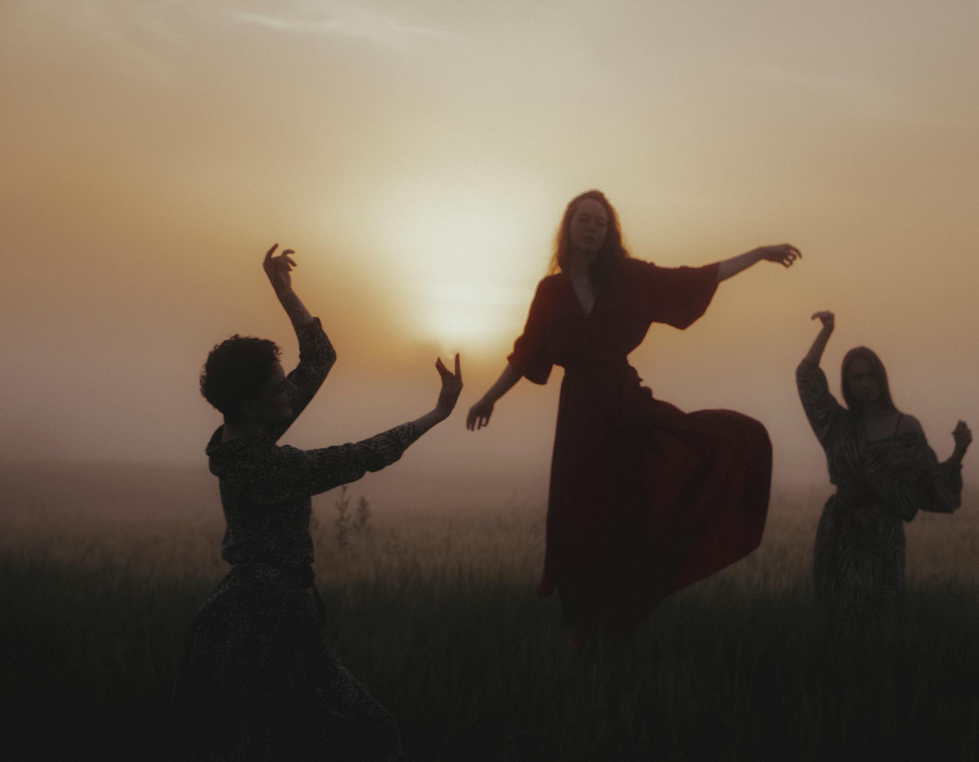 Three women dancing gracefully in a misty field at sunrise, creating an ethereal backlit silhouette.