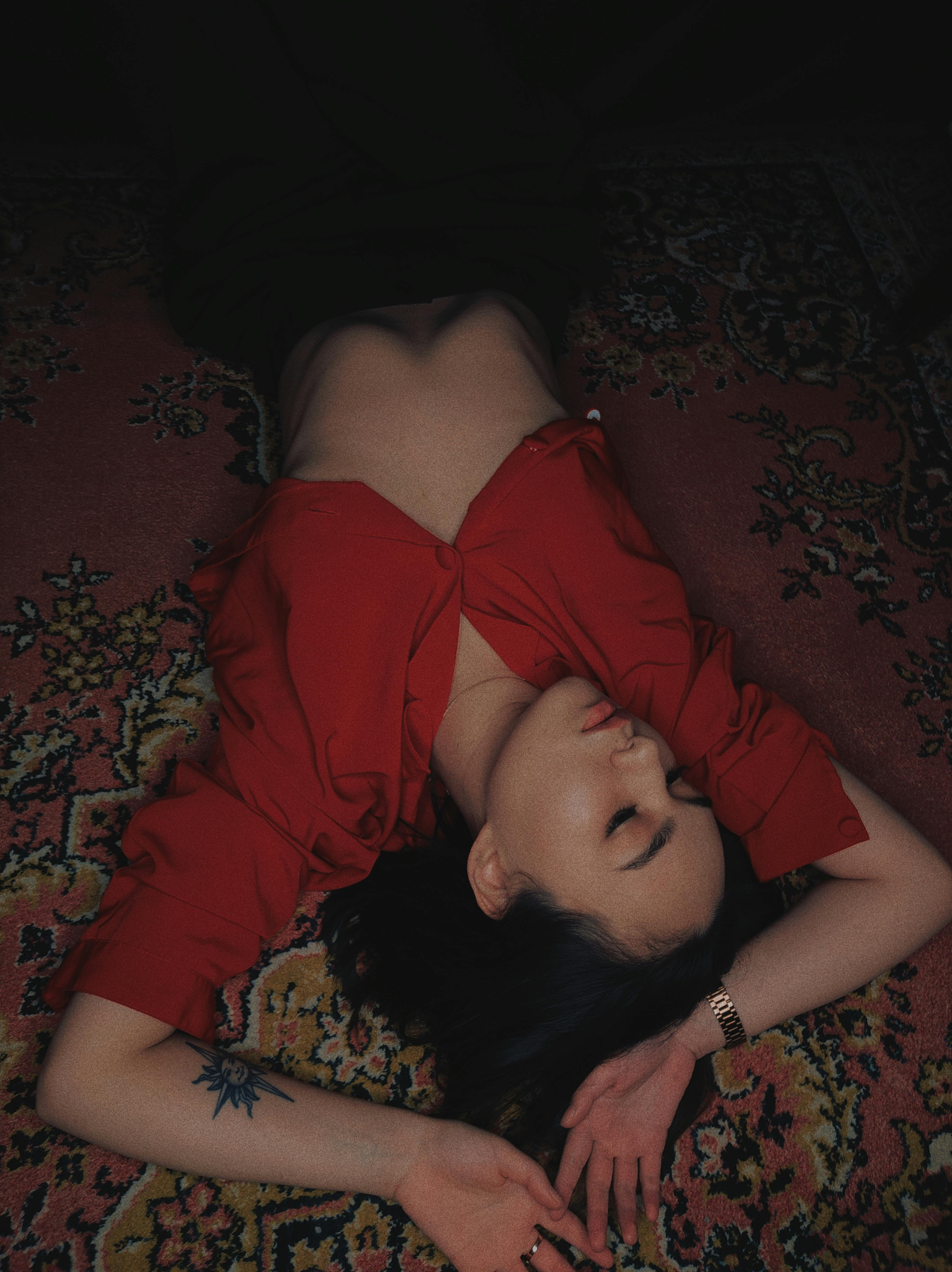 A woman with a tattoo relaxes on a patterned rug, wearing a red crop top, eyes closed and arms raised.