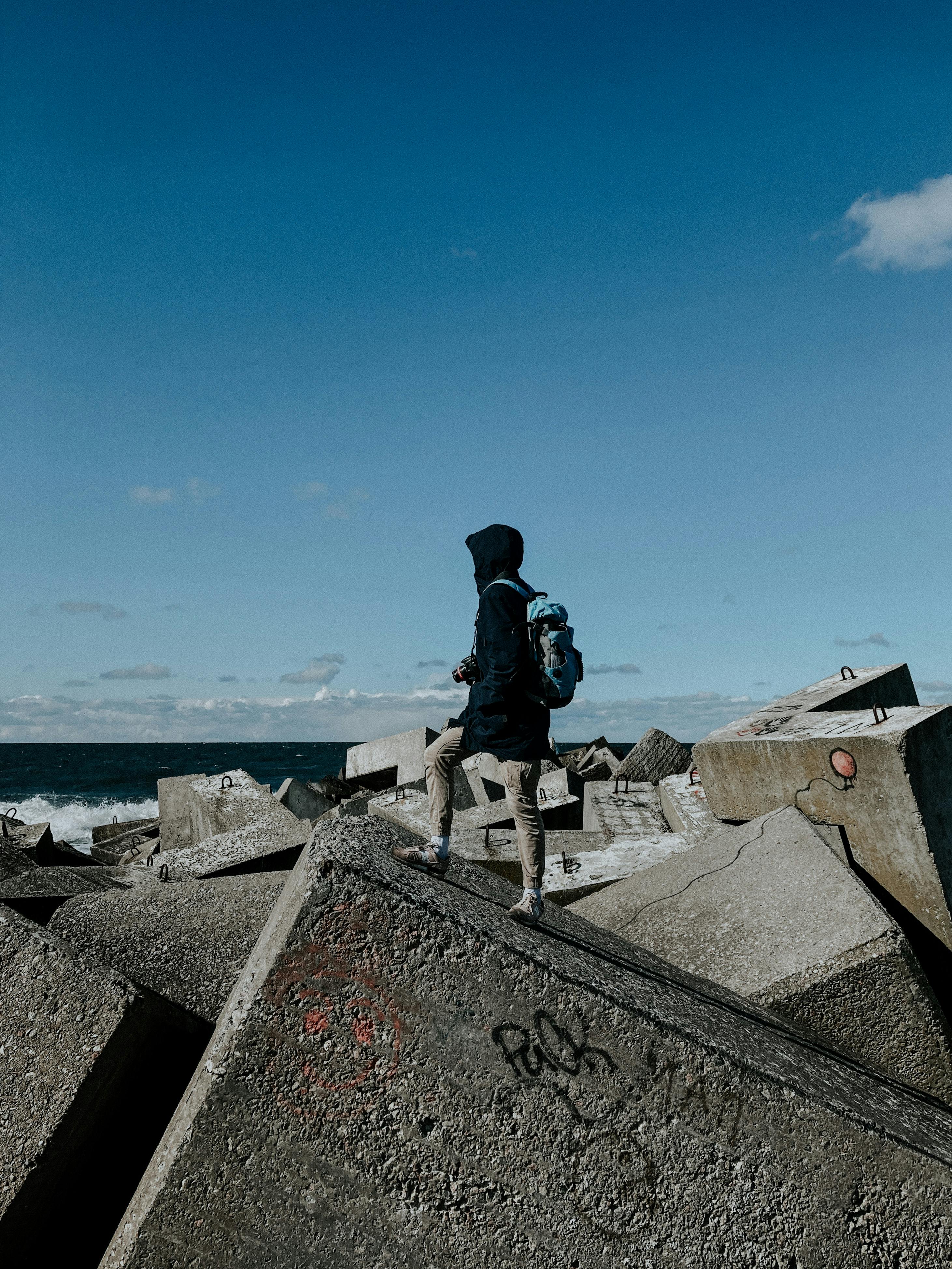 An individual standing on rocky concrete blocks by the sea under a clear blue sky.